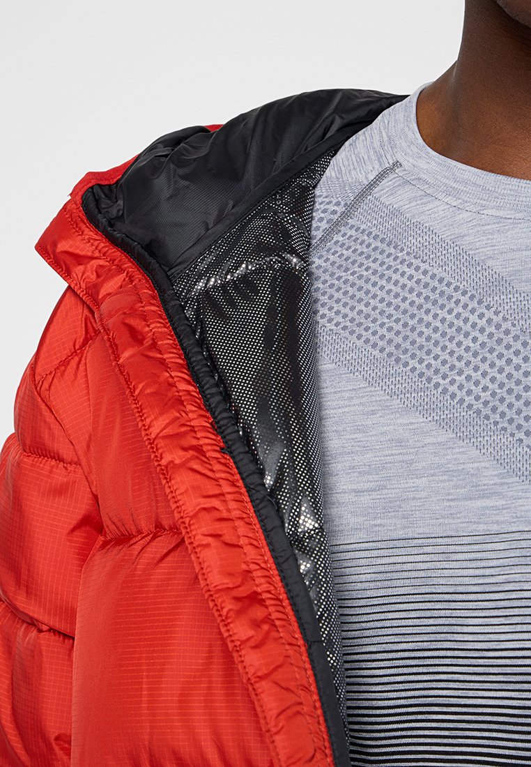 Chaqueta acolchada roja con superficie texturizada, forro negro y acentos metálicos, llevada sobre una camiseta gris con patrones a rayas.