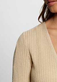 Beige värillinen ribbineulottu paita V-pääntiellä, jossa on teksturoitu kangas ja metallinhohtoiset langat korostuksena. Lähikuva olkapäästä ja pääntiestä.