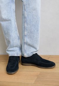 Pantofi negri din suede cu un finisaj neted, design cu șireturi, vârf rotunjit și talpă din cauciuc, asortați cu pantaloni din blugi bleu deschis.