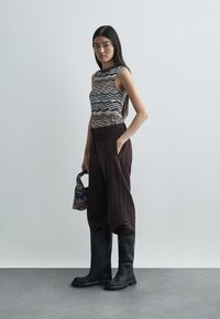 Bruna byxorna med zigzag-mönster i vid modell, en åtsittande tanktop med en vågig multicolor-design och svarta knähöga stövlar. En liten mönstrad handväska.