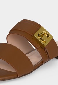 Καφέ δερμάτινο σανδάλι τύπου slide με δύο λουριά και χρυσή τετράγωνη αγκράφα με διακοσμητικό σχεδιασμό που διαπλέκεται.