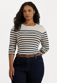 Donna con i capelli ricci che indossa una maglietta a righe crema e blu navy a maniche lunghe, jeans scuri, una cintura marrone con fibbia dorata e orecchini a cerchio dorati.