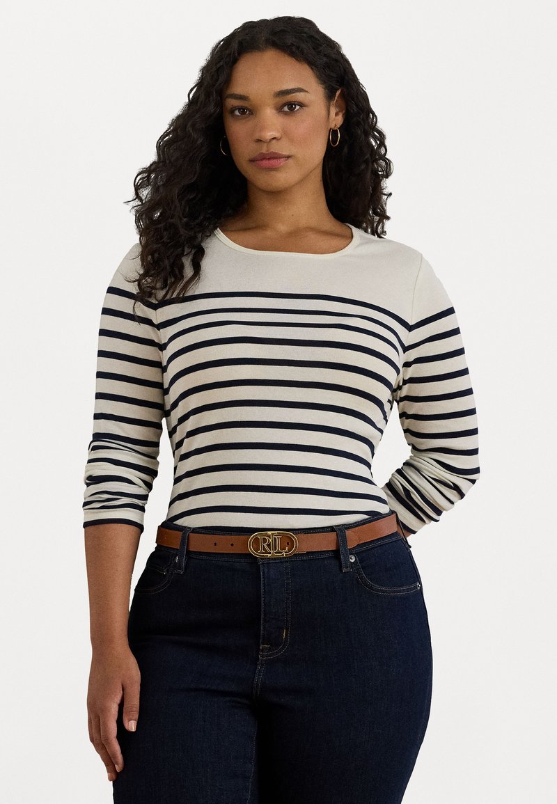 Donna con i capelli ricci che indossa una maglietta a righe crema e blu navy a maniche lunghe, jeans scuri, una cintura marrone con fibbia dorata e orecchini a cerchio dorati.