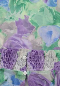 Tissu fleuri violet, vert, bleu et blanc avec une taille élastique froncée et deux petits boutons blancs cousus.