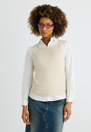 Vrouw met krullend haar, rode bril, beige mouwloze top over een wit shirt, en een blauwe denim rok tegen een effen achtergrond.