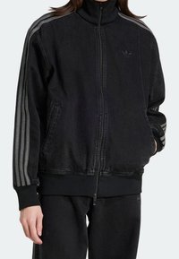 Svart zip-up jacka i denim, med hög krage, två sidofickor och randiga detaljer i grått på axlarna.