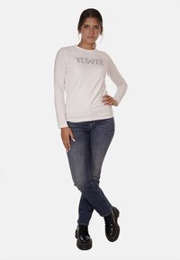 Maglietta bianca a maniche lunghe con la scritta "YES-ZEE" stampata in argento, abbinata a jeans blu e stivaletti neri con lacci. Il modello sta in piedi con le braccia incrociate.