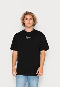 Svart kortärmad t-shirt i bomull med en vit broderad logotyp på vänster bröst. Modellen är matchad med ljusblå jeans. Neutral bakgrund.