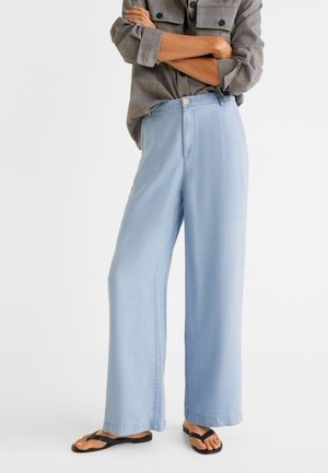 Osoba v světle modrých kalhotách s širokými nohavicemi, šedé kostkované košili s dlouhým rukávem, který má vyhrnutý, a černých žabkách stojící se zařízenýma rukama.