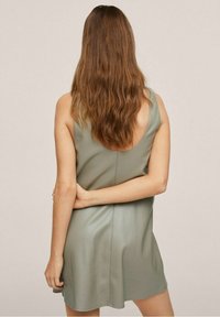 Vrouw met lang bruin haar, gekleed in een mouwloze, lichtgroene jurk, staande met een gebogen arm achter haar rug.
