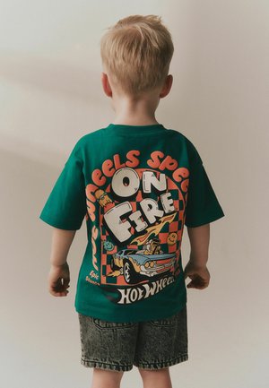 Child with short blond hair facing away, wearing green Hot Wheels t-shirt with colorful car and "On Fire" text, and denim shorts.