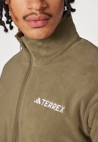 Fleece zip-up jacket in olive green with a high collar. Features white "TERREX" logo and soft texture, designed for comfort.