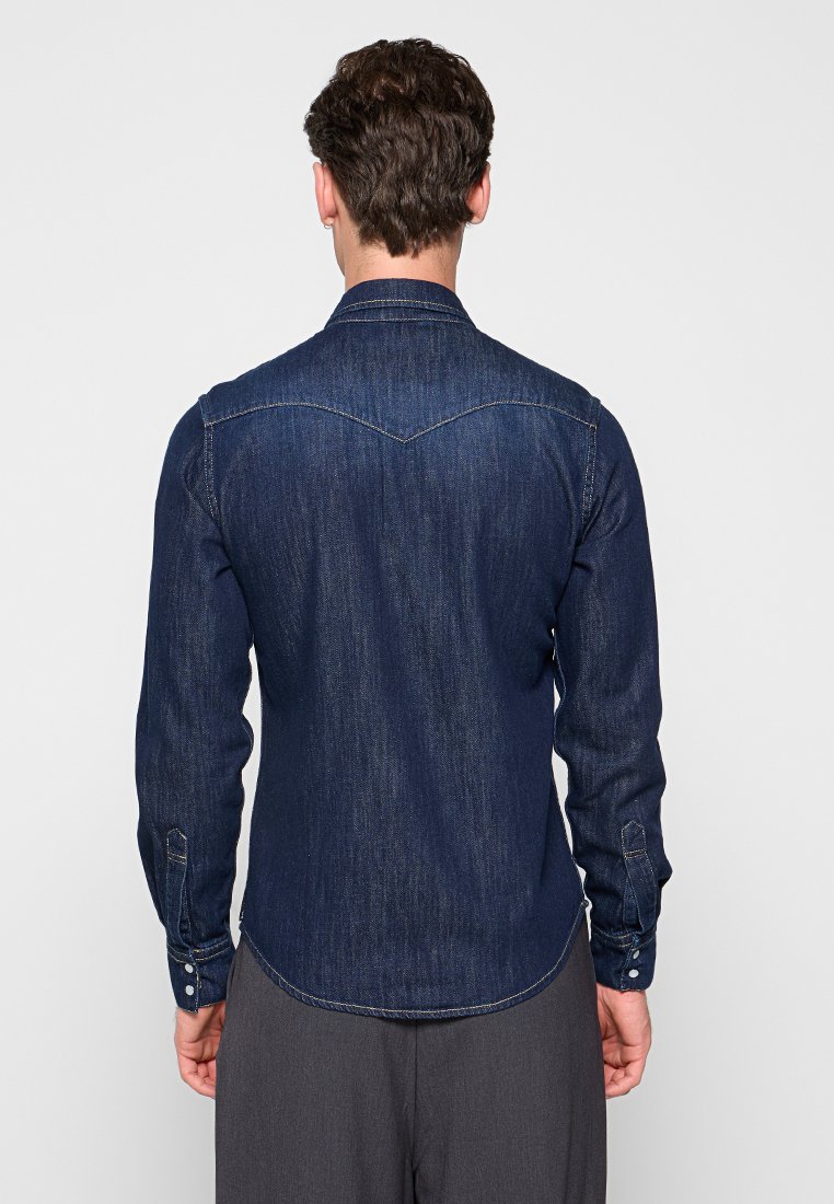 Dunkelblaue Jeanshemd mit Yoke-Design, langen Ärmeln, Knopfmanschetten und einem abgerundeten Saum. Der Stoff hat eine strukturierte Oberfläche und sichtbare Nähte.