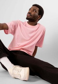Lyserød bomulds T-shirt med korte ærmer, parret med sorte bukser og hvide sneakers, siddende mod en grå baggrund.