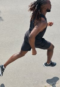 Svart ärmlös atletisk topp och shorts, springer på asfalt, bär svarta och vita sneakers, muskulös byggnad, i solljus.