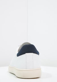 Baskets en cuir blanc avec un onglet de talon en tissu bleu marine, une semelle en caoutchouc beige, une surface lisse et un design minimaliste. Vue arrière mettant en avant les détails de la couture.