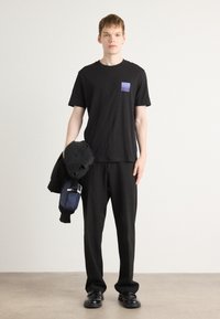 T-shirt noir avec un patch logo violet, associé à un pantalon noir et des chaussures noires. Le modèle tient une veste noire et un sac bleu marine avec des accents argentés.