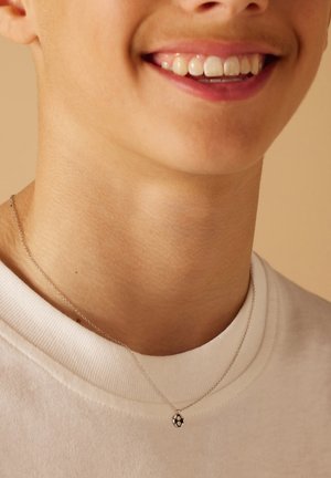 Zilveren ketting met een kleine ronde hanger met een honingraatpatroon, gedragen boven een witte t-shirt met ronde hals.