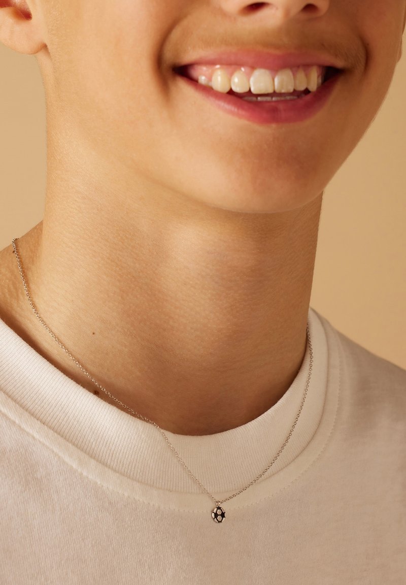 Zilveren ketting met een kleine ronde hanger met een honingraatpatroon, gedragen boven een witte t-shirt met ronde hals.