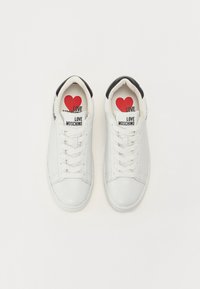 Baltas ādas sporta apavi ar melniem papēža akcentiem, plakano auklu un "Love Moschino" uzlīmēm. Sarkanā sirds grafika uz zolēm.