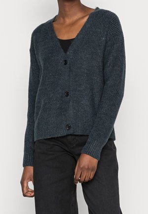 Tmavě šedý žebrovaný kardigan se třemi černými knoflíky, nošený přes černý top s dlouhými rukávy, v kombinaci s černými kalhotami.