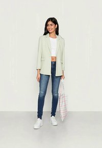 Blazer verde claro com lapelas, bolsos frontais, top branco cropped, jeans escuros e sapatilhas brancas. A segurar uma mala de ombro em padrão xadrez rosa.
