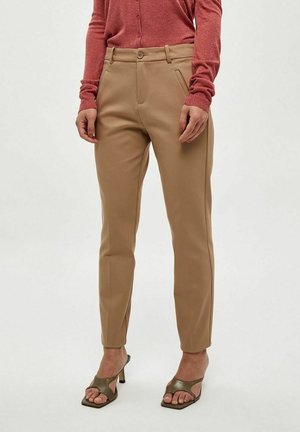 Personne portant un pantalon beige sur mesure longueur cheville, un haut rouge à manches longues et des sandales à talons ouvertes olive.