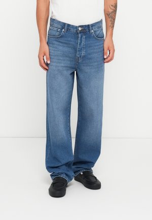 Man in een losse blauwe spijkerbroek en zwarte schoenen, staande tegen een effen witte achtergrond.