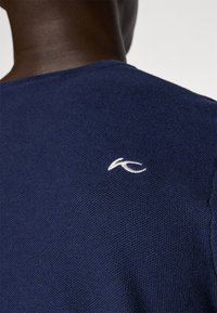 Sweater de malha texturizada azul-marinho com logótipo branco na parte de trás da gola. Apresenta um decote redondo e mangas curtas. O material parece suave e respirável.