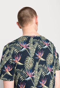 Person med kort hår som vänder bort, iklädd en mörk skjorta mönstrad med stora tropiska löv och orange-rosa fågelparadisblommor.