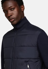 Chaleco azul marino con cremallera en la parte delantera, con frente acolchado, cuello de canalé y bolsillos laterales con cremallera. Fabricado con un tejido suave y ligero, de diseño limpio.