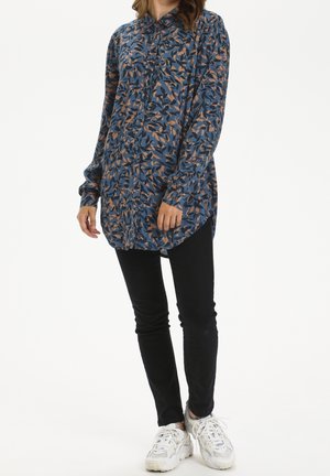 Woman wearing blue and brown leaf-patterned long-sleeve button-up shirt, black slim pants, and white sneakers, standing against white background.