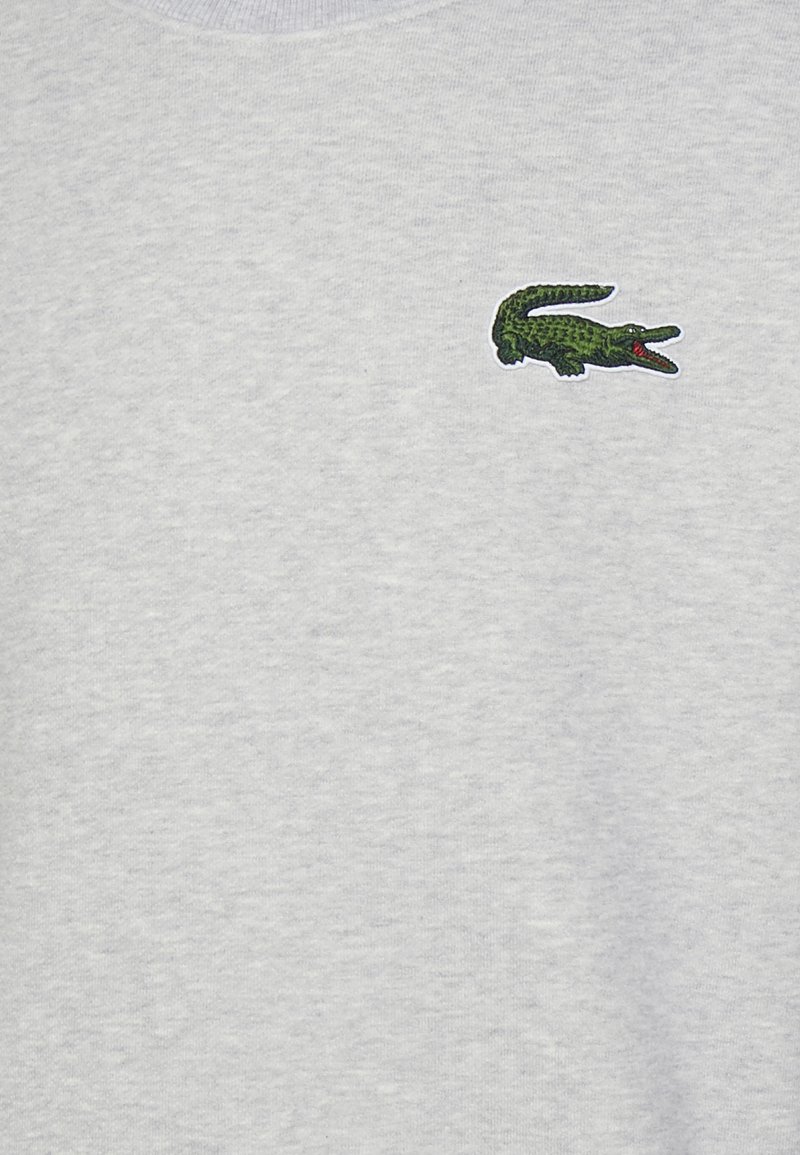Szary sweatshirt z teksturowanej tkaniny, z prążkowanym dekoltem i wyhaftowanym zielonym logo krokodyla na lewej piersi.