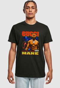 Sort bomulds T-shirt med et grafisk print af en muskuløs mand med teksten "GUCCI MANE" i fede røde og gule bogstaver.