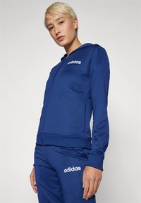 Zila sporta zip-up jaka no gludas auduma, ar kapuci un ribotām aprocēm, ar izšūtu balto adidas logo uz krūtīm.