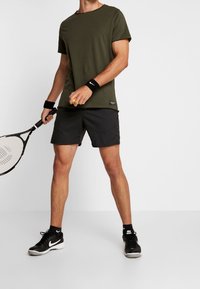 Olivově zelené tričko a černé kraťasy. Bavlněná směsová tkanina, kulatý výstřih, přiléhavý střih. Drží tenisovou raketu s černými zápěstními pásky a tenisovým míčkem.