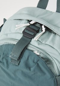 Grön ryggsäck med mjukt tyg, justerbara remmar, en dragkedjeficka och svarta detaljer med vita dragsnören.