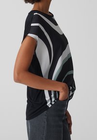 Osoba ubrana w bezrękawnik w czarno-białe-zielone wzory, z jedną ręką w kieszeni ciemnoszarych spodni, na jednolitym tle.