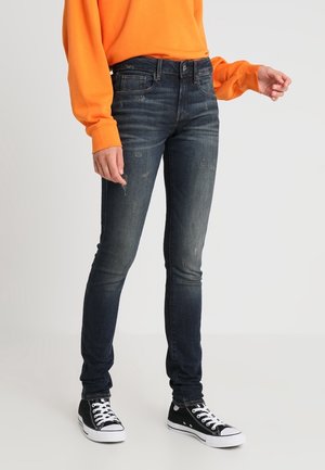 Person trägt dunkelblaue Slim-Fit-Jeans, schwarz-weiße Sneaker und ein orangefarbenes Langarmshirt, steht vor hellem Hintergrund.