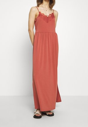 Femme portant une robe rouille sans manches, longue jusqu'aux chevilles, ornée d'un galon en dentelle au niveau de l'encolure, des sandales beiges et des colliers superposés.