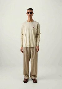 Man in een beige overhemd met lange mouwen en klein logo, losse beige broek, bruine schoenen en een zonnebril, staande tegen een effen lichte achtergrond.