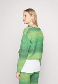 Suéter cropped em ombre verde com mangas longas, textura malha e decote largo, combinado com um top branco e leggings verdes a condizer.