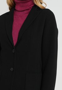 Černý blazer se dvěma knoflíky, klopovými límci a dvěma předními kapsami, navrstvený na přiléhavém fialovém roláku.