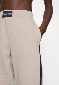 Beżowe spodnie dresowe Calvin Klein z prążkowanym pasem, kieszeniami po bokach oraz czarnymi bocznymi wstawkami z logo. Gładki materiał bawełniany.