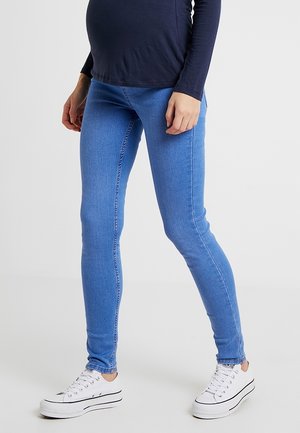 Person wearing blue skinny jeans, white platform sneakers, and a navy long-sleeve top standing against a plain background.
