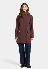 Bordeaux waterdichte jas met een capuchon, voorzien van twee voorkoten, drukknoopsluitingen en een rechte snit, gecombineerd met blauwe jeans en zwarte laarzen.