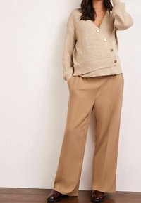 Pull beige côtelé avec un col en V et de gros boutons, associé à un pantalon large taille haute en khaki. Des chaussures marron complètent la tenue.