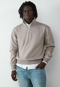 Beige Halb-Reißverschluss-Sweatshirt mit glatter Textur, gerippten Bündchen und Saum, getragen über einem weißen Hemd, kombiniert mit hellblauen Jeans.