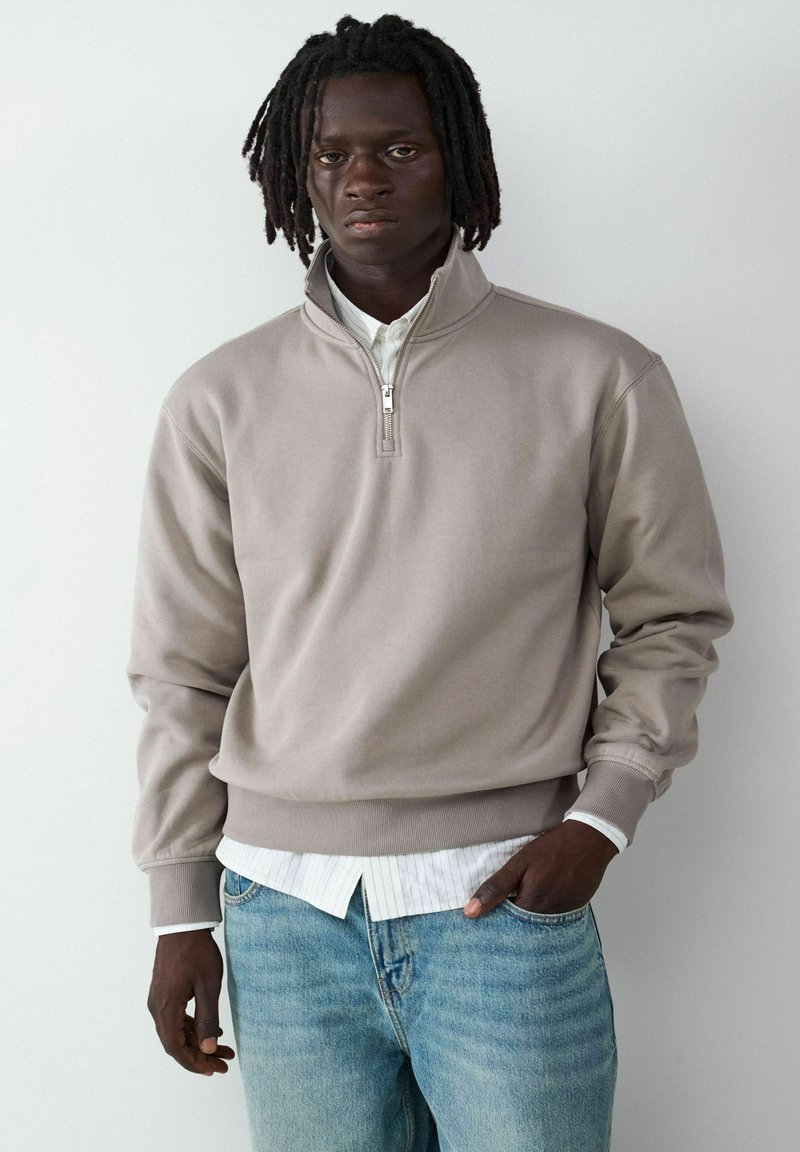 Beige Halb-Reißverschluss-Sweatshirt mit glatter Textur, gerippten Bündchen und Saum, getragen über einem weißen Hemd, kombiniert mit hellblauen Jeans.