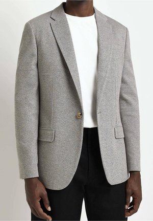 Mann trägt einen grauen, strukturierten Blazer mit einem einzelnen Knopf, weißes Hemd und schwarze Hose vor einem schlichten, hellen Hintergrund.